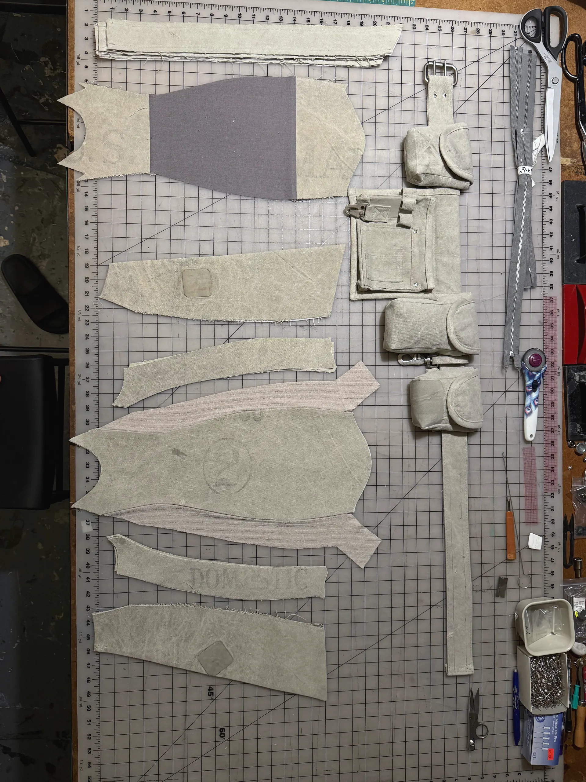 An overhead view of a cutting table holding the cut canvas pieces of a garment alongside a finished utility harness: a long strap with a metal buckle carrying four snap-flap pockets and a flat zipped pouch. Shears, a metal rule, a rotary cutter, an awl and an open tin of pins are ranged down the right-hand side.