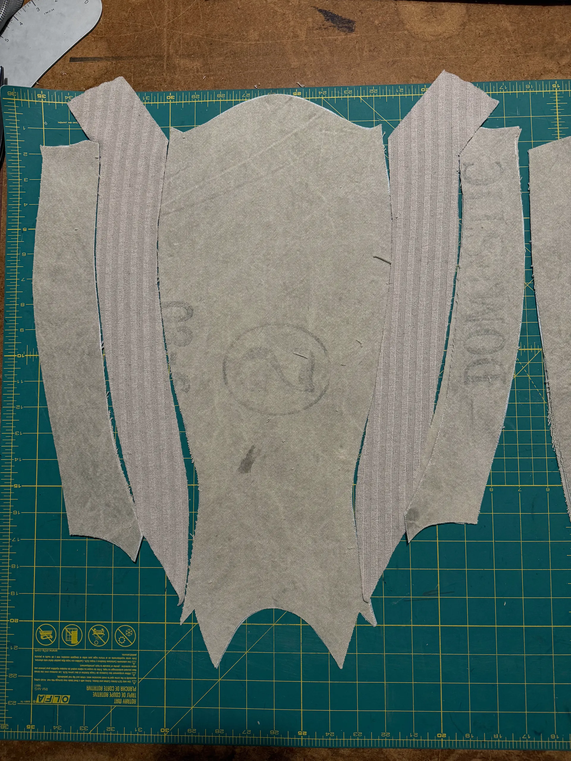 Cut pattern pieces for a vest laid out on a green gridded cutting mat: a wide centre panel with a scalloped, pointed hem flanked by narrow curved side and facing pieces. The pale canvas carries faded printed lettering and a circled mark left over from its previous life as printed cloth.