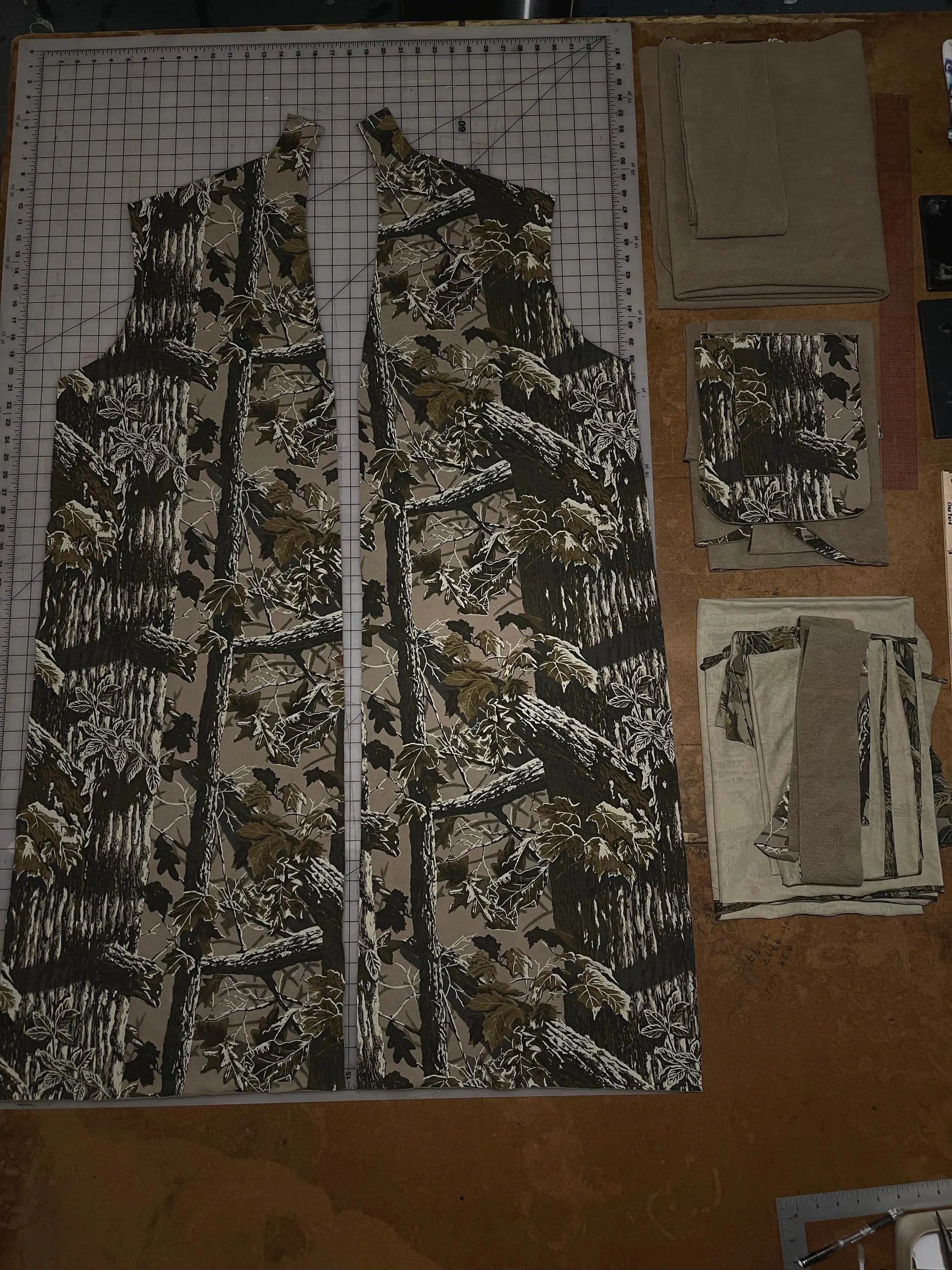 Two long cut panels of a tree-bark and leaf camouflage print lie mirrored on a gridded cutting mat, forming the fronts of a full-length coat. Stacked bundles of matching camouflage and plain olive pieces, including pockets and facings, are sorted on the brown table beside them.