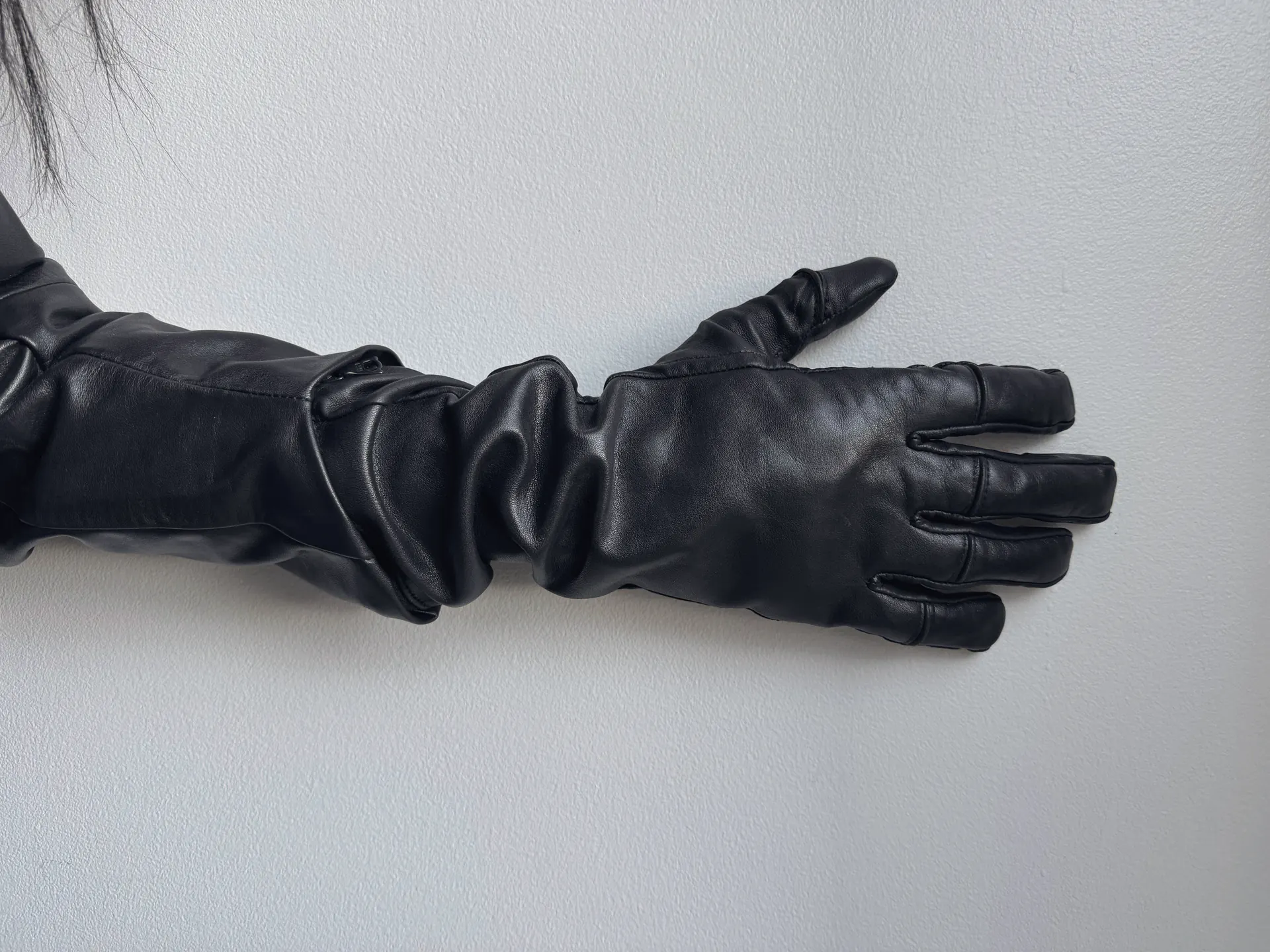 Black leather glove at the end of a black leather sleeve, arm extended against a pale grey wall