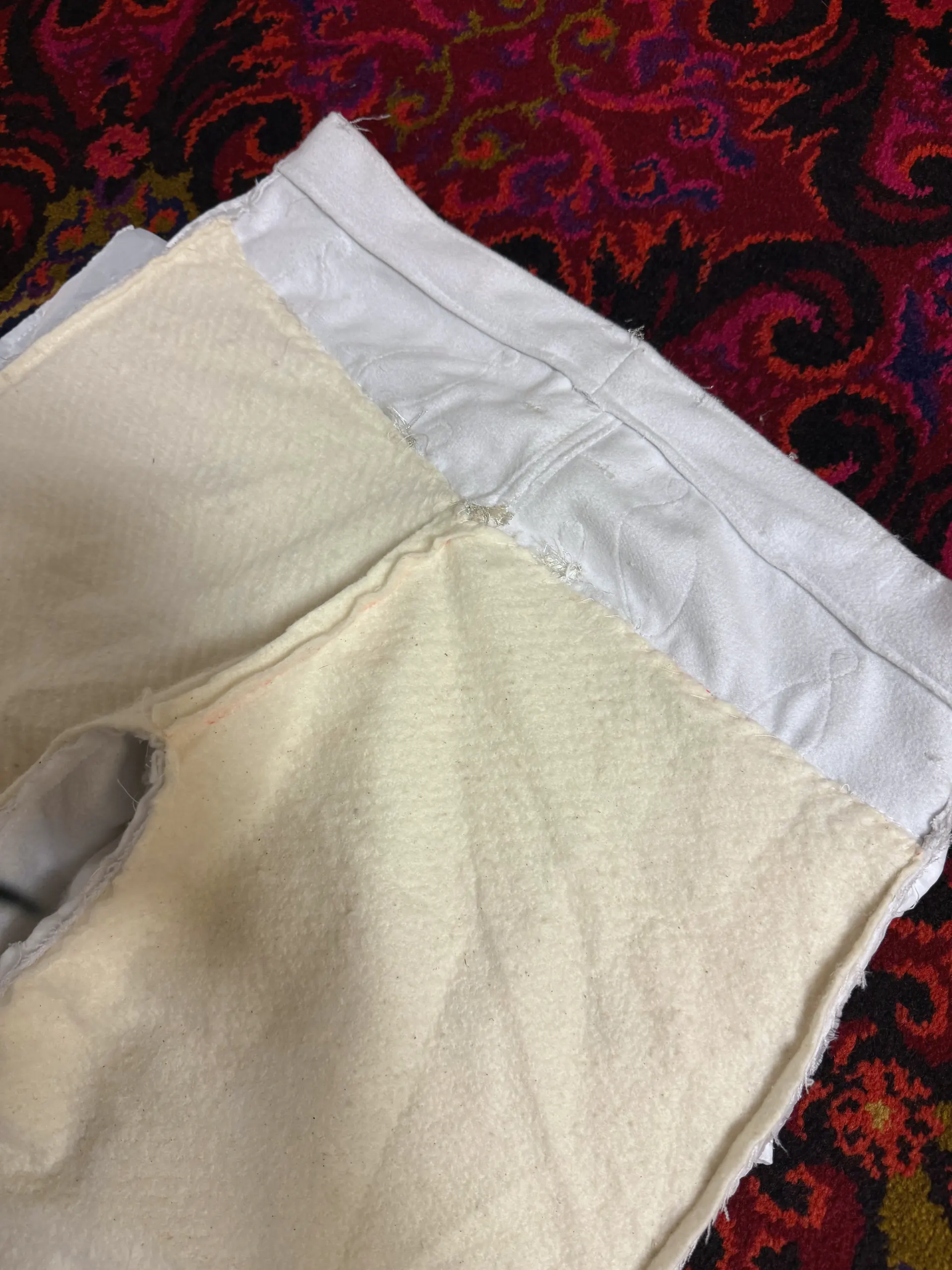 A close detail of the waistband corner of a pair of white trousers, where a cream felted inner layer meets a smooth pale outer shell. The join is sewn with a raw seam allowance left standing on the face of the cloth, and a small hand-worked bar tack secures the centre seam.