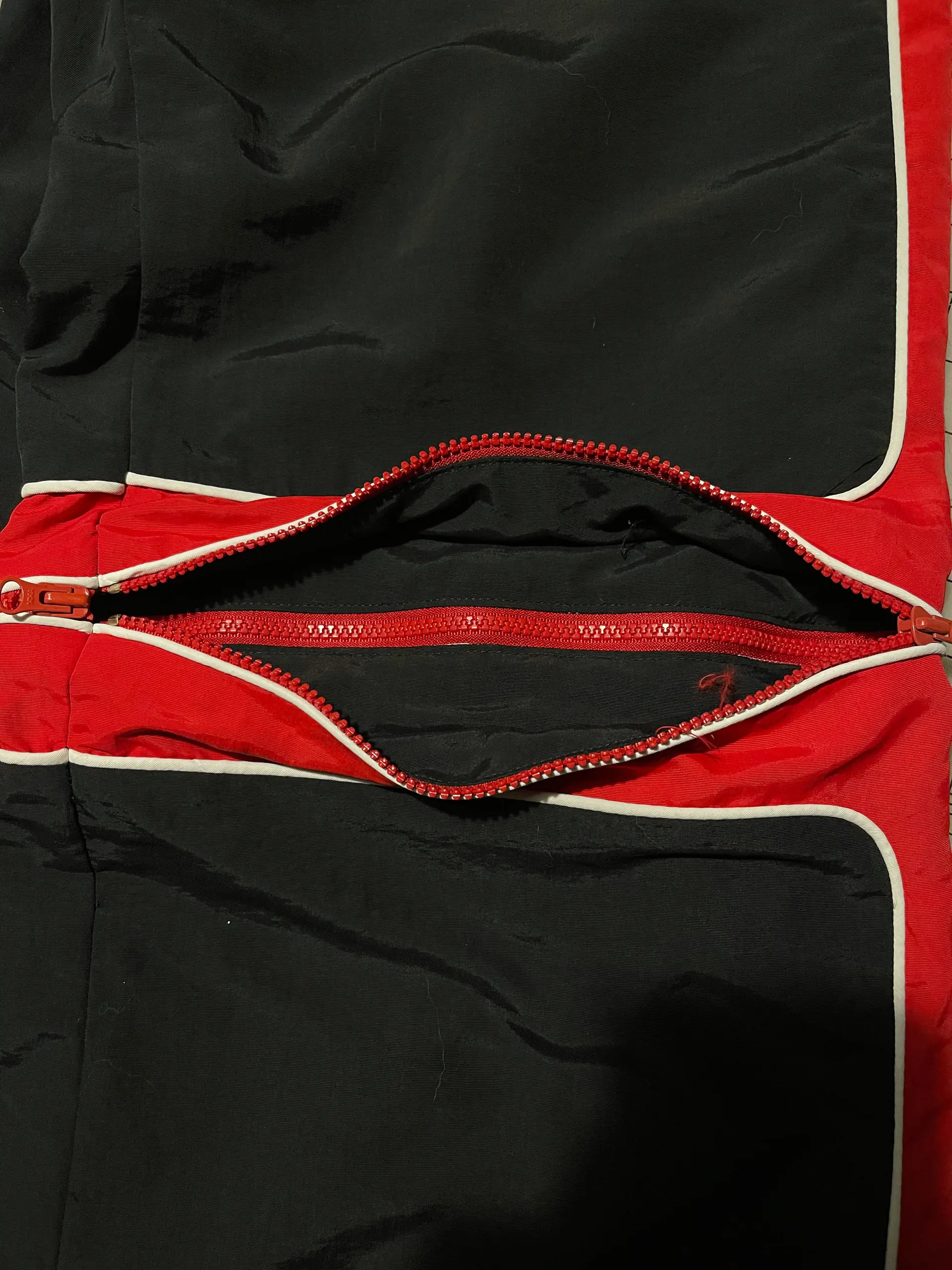 The knee zip on the black trousers pulled part-way open, the two red coil tapes splayed apart to show the dark inside faces of the leg. White piping edges the red band above and below the opening, with loose red thread ends left at the seam.