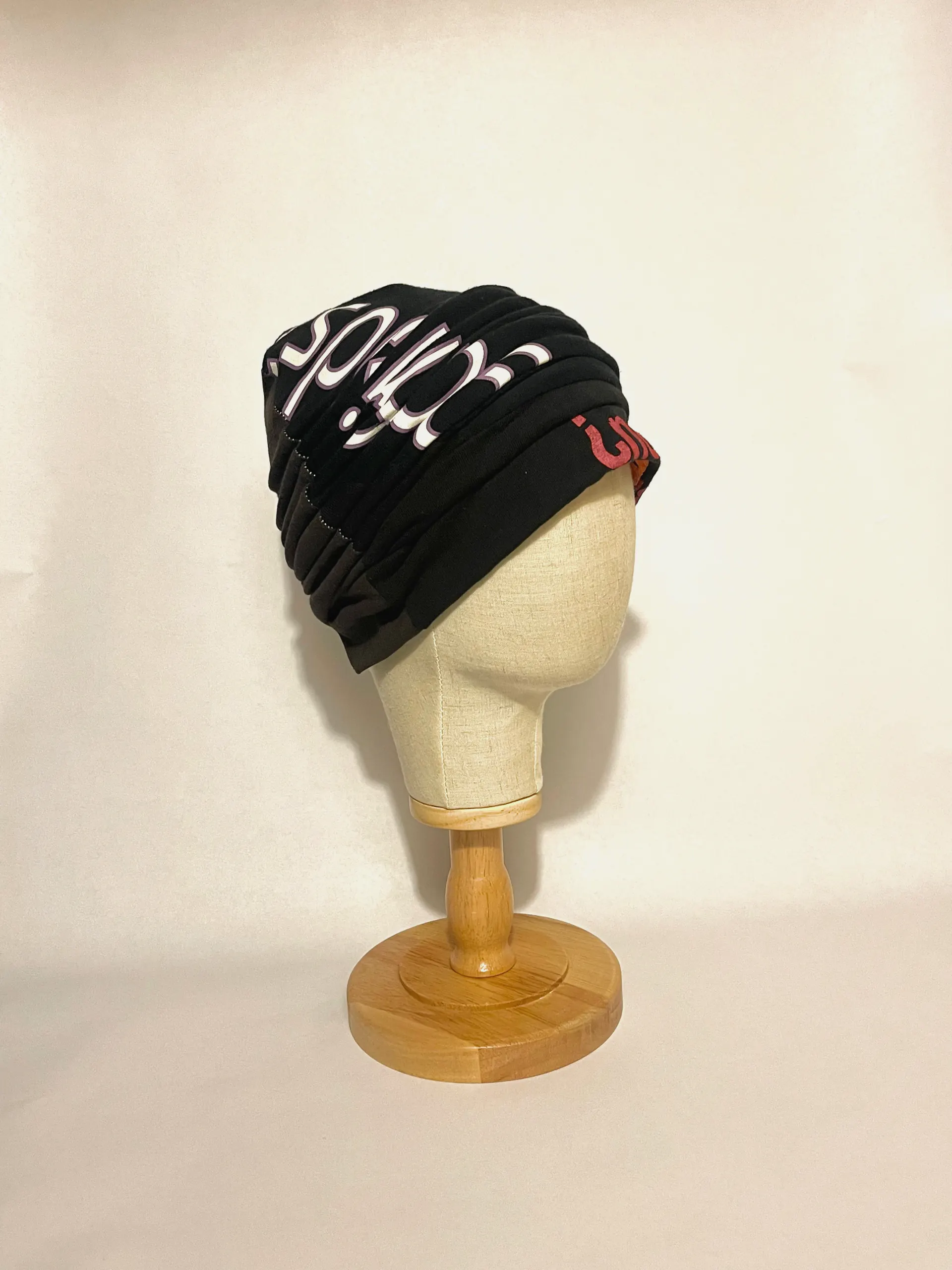Black pleated jersey head wrap on a linen head form, opposite three-quarter view showing the printed lettering