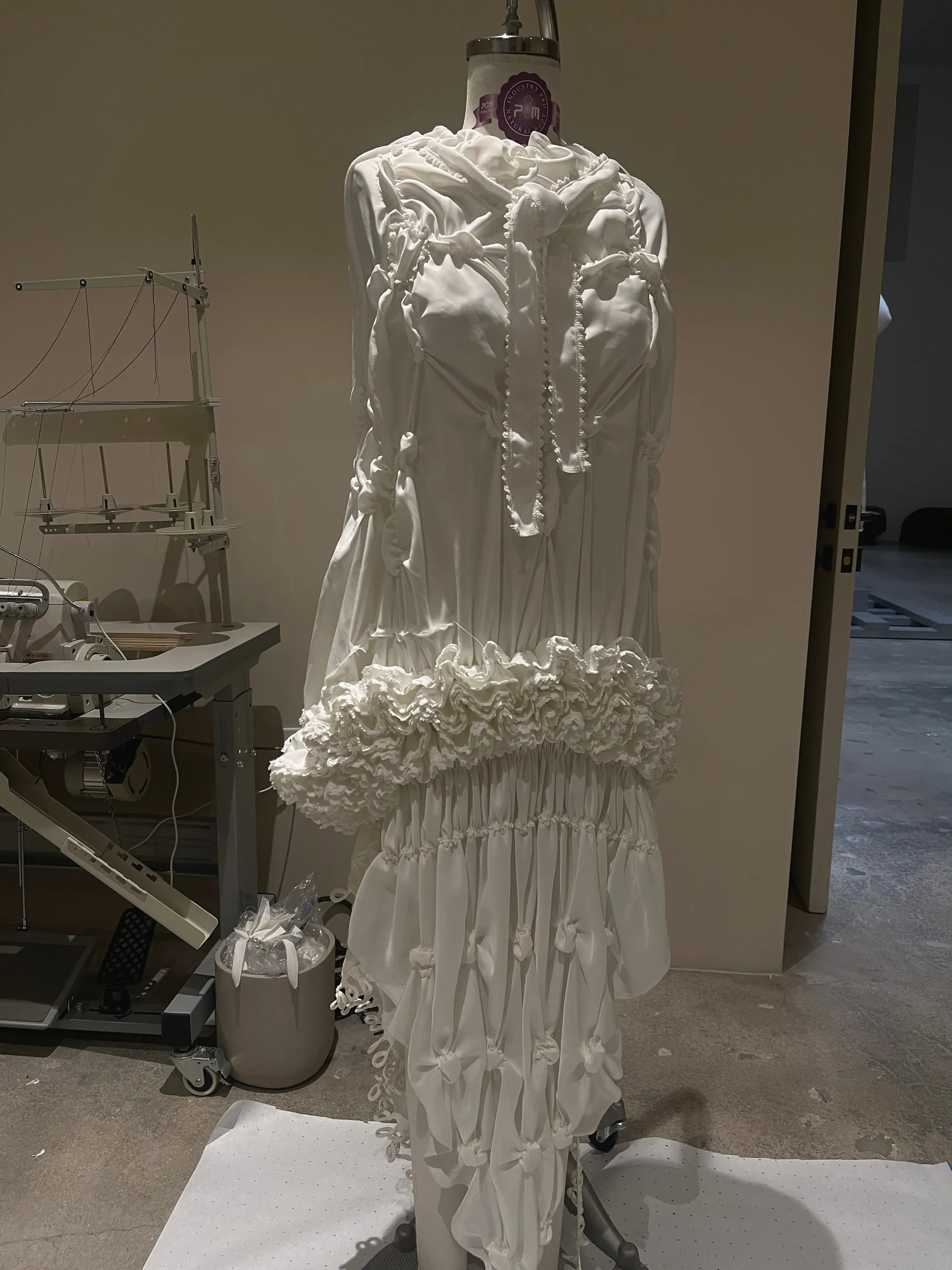 White long-sleeved gown with a shirred bodice, knotted bow trims, a ruffled band at the hip and a tiered gathered skirt, on a dress form in the studio.