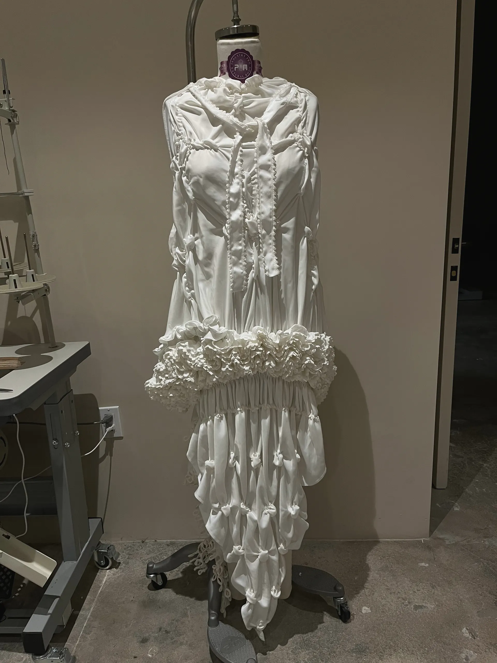 White long-sleeved dress on a dress form: ruched bodice with self-covered buttons, a ruffled band at the hip and a gathered, knotted skirt with a train.