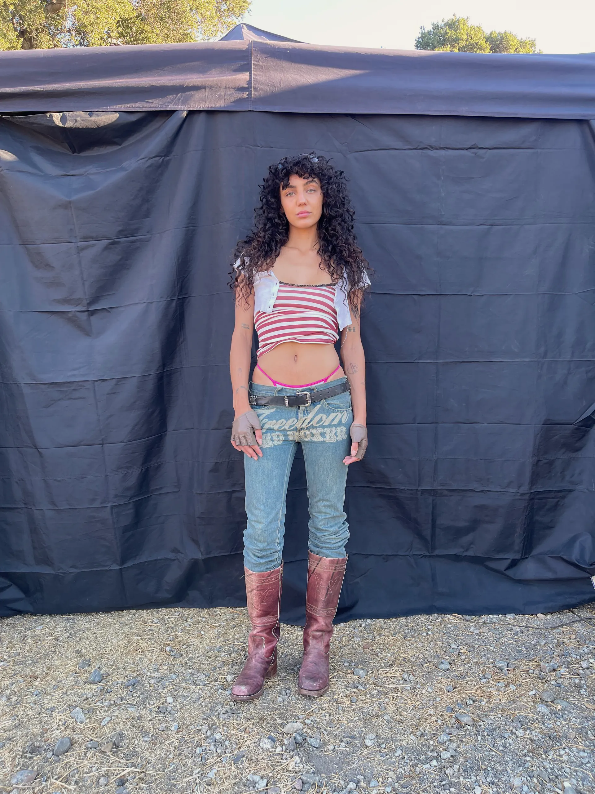 A model stands against a black tarpaulin backdrop on gravel, wearing a cropped red-and-white striped top with pale shoulder panels over very low-rise faded jeans carrying a printed graphic across the front, finished with fingerless gloves and tall oxblood boots.