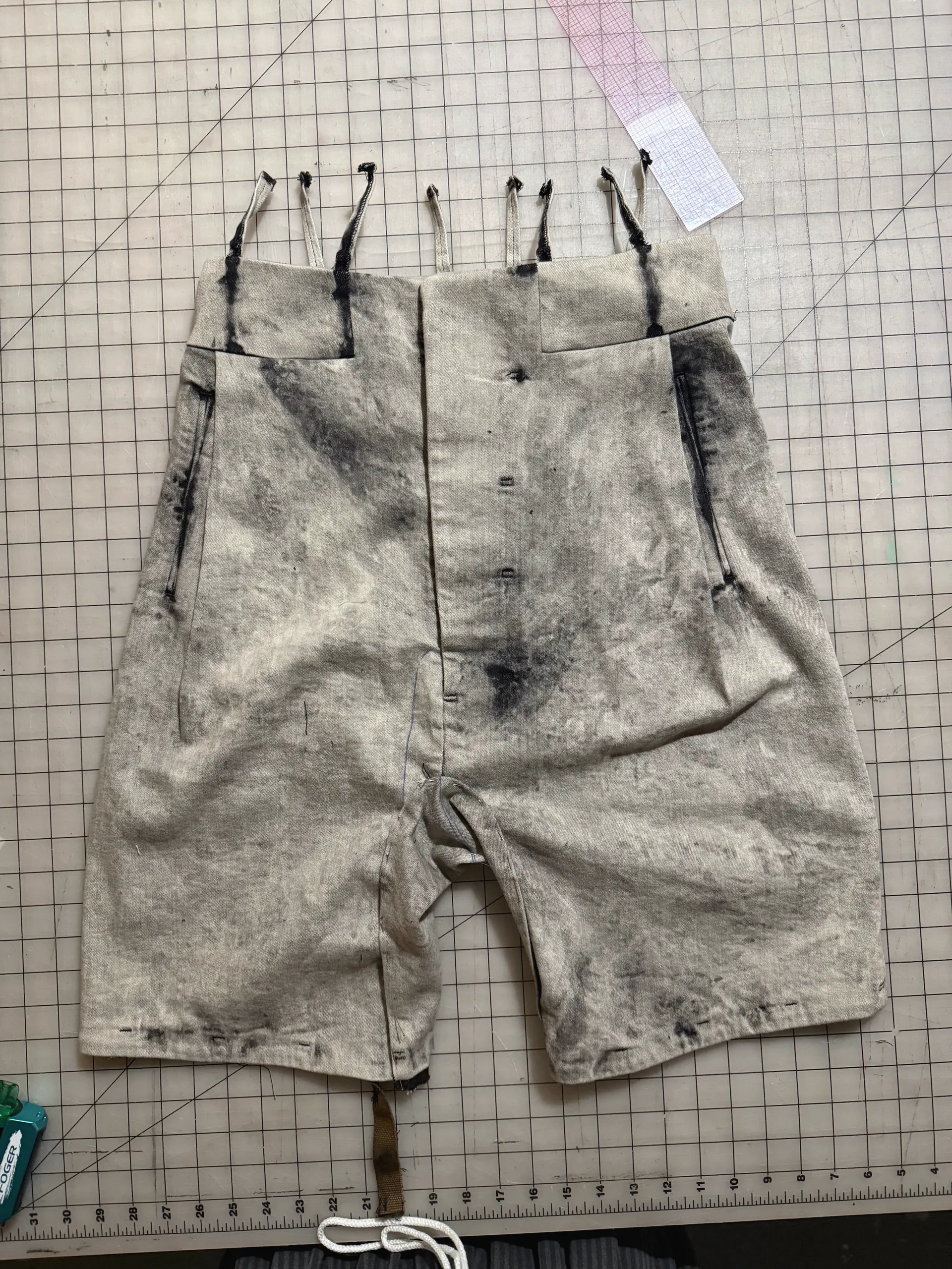 Mottled off-white and grey hand-dyed cotton shorts with frayed waistband tabs and button fly, flat on a cutting mat.