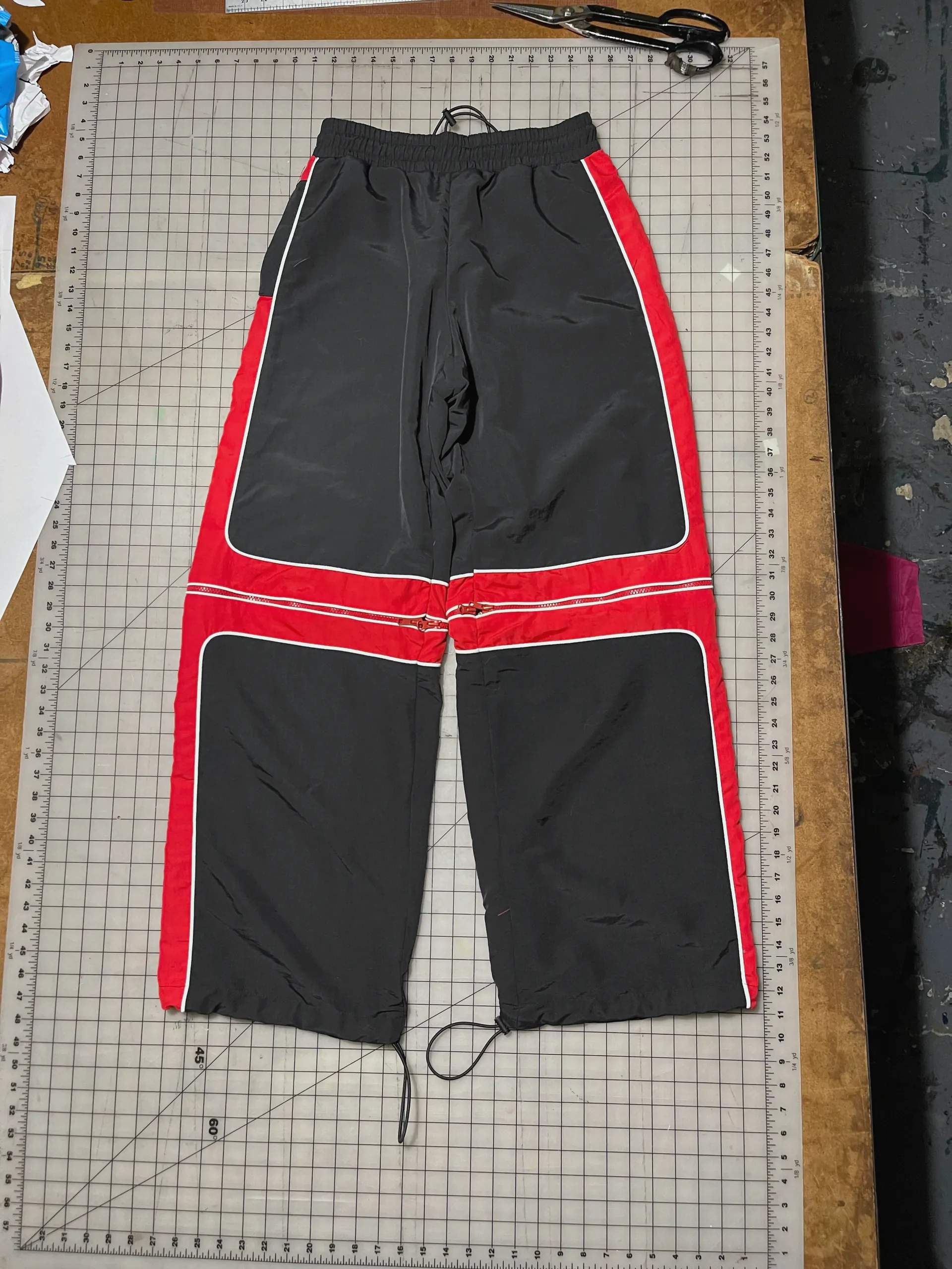 Wide-legged black trousers laid face-up on a gridded cutting mat, with red side panels and a red band circling each leg at the knee. Twin zips run through the knee bands so the lower legs detach, every colour break edged in white piping, an elasticated drawcord waist at the top and cord ties at the hems.