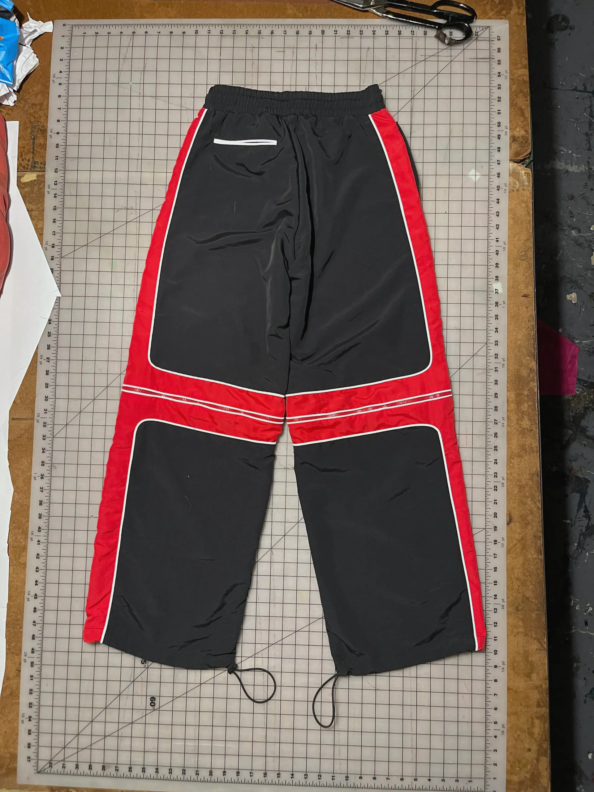 The wide black trousers turned over on the cutting mat to show the back, with red side panels running the full length of the legs, the red knee bands and their zips carrying round, a welt pocket set below the elasticated waistband and cord ties at each hem.