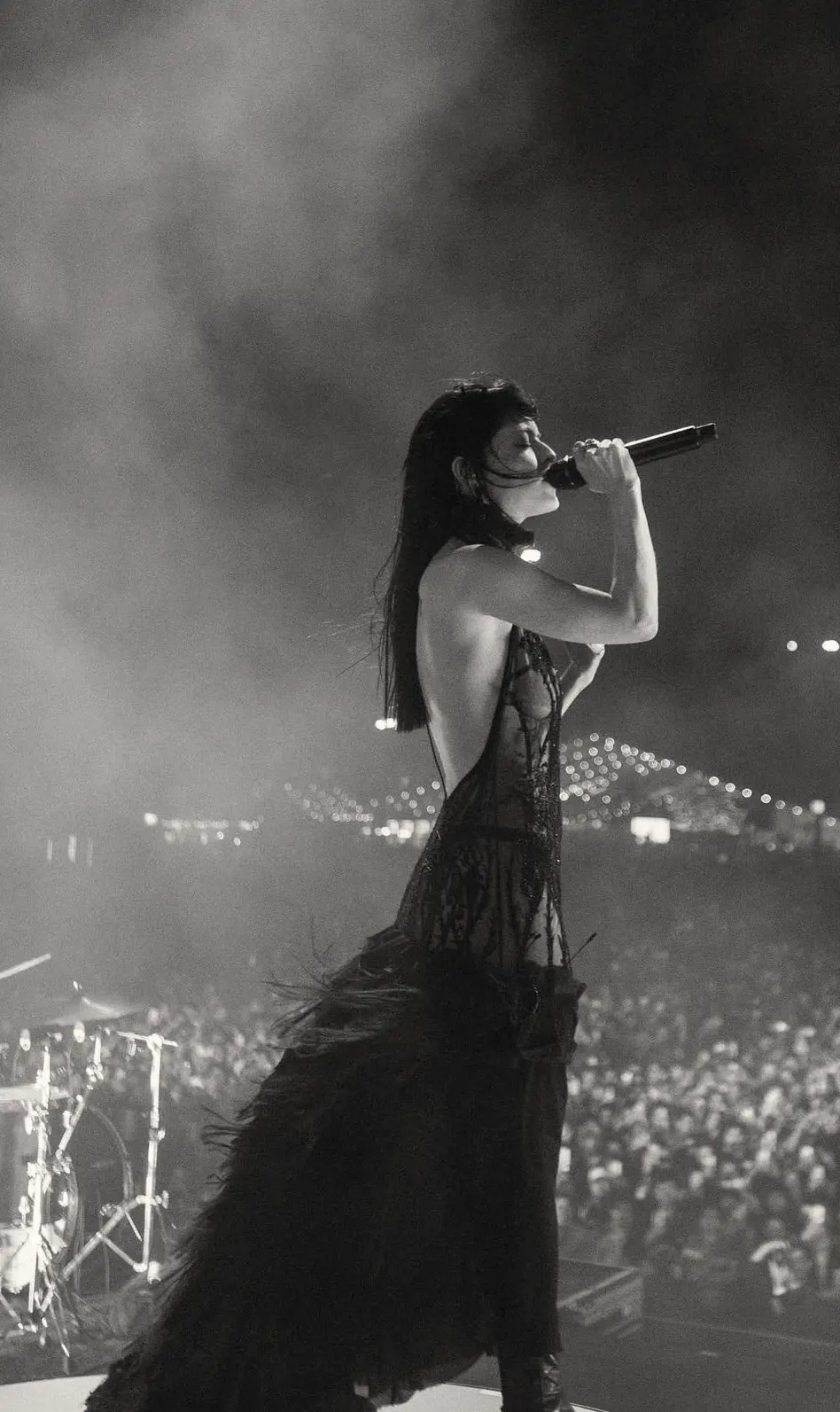 Black-and-white photograph of a performer singing into a handheld microphone on stage before a large outdoor crowd, wearing a black open-back gown with a lace bodice and a long tiered ruffled skirt.